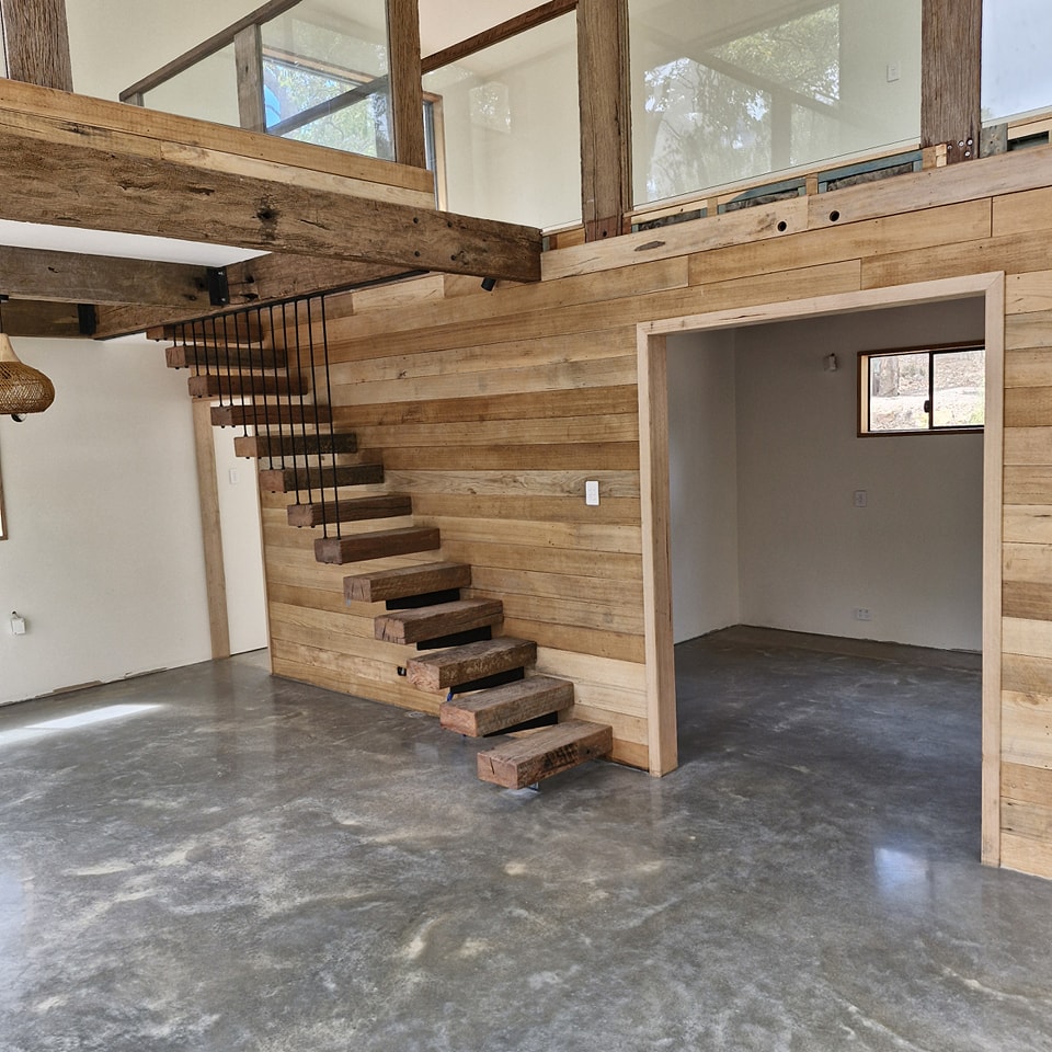481003176_122120698520695867_7053531817514354707_n Modern interior with polished concrete floor, floating wooden staircase, and wood-panelled walls leading to an open room and loft area.
