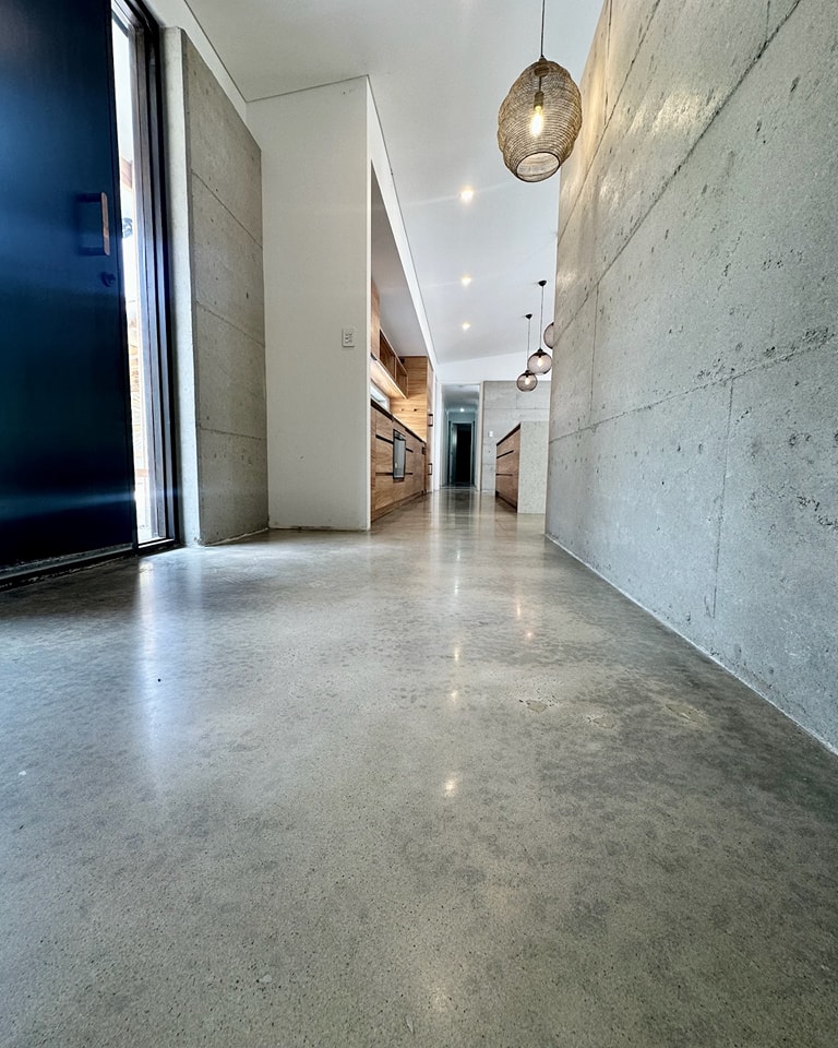 Full Exposure Finish A modern hallway with polished concrete floors, concrete walls, pendant lights, and wooden accents, leading to an open door at the far end.