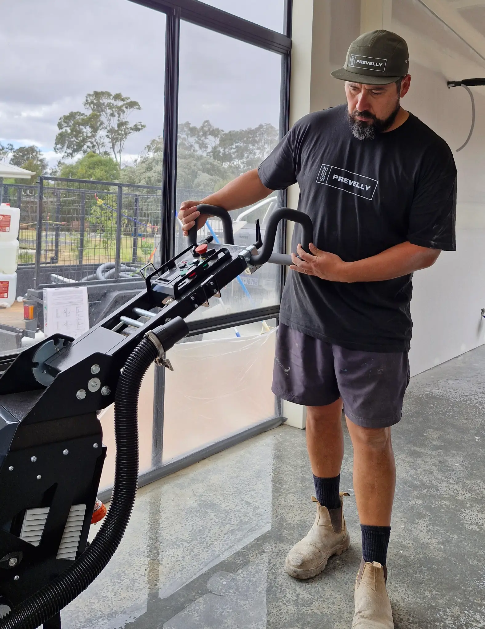A man wearing a Prevailly t-shirt and cap operates a floor polishing machine inside a building under construction.